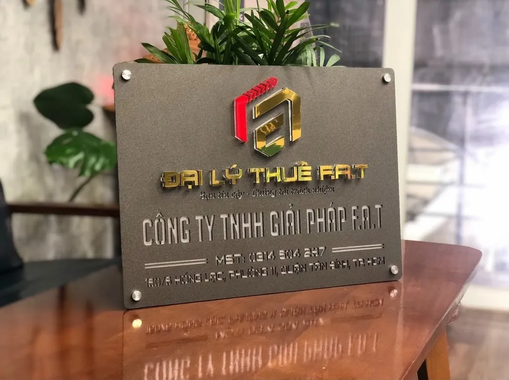 Bảng hiệu công ty inox xám khói logo nổi 3D vàng đỏ bạc nhìn góc nghiêng 45 độ trên mặt bàn gỗ