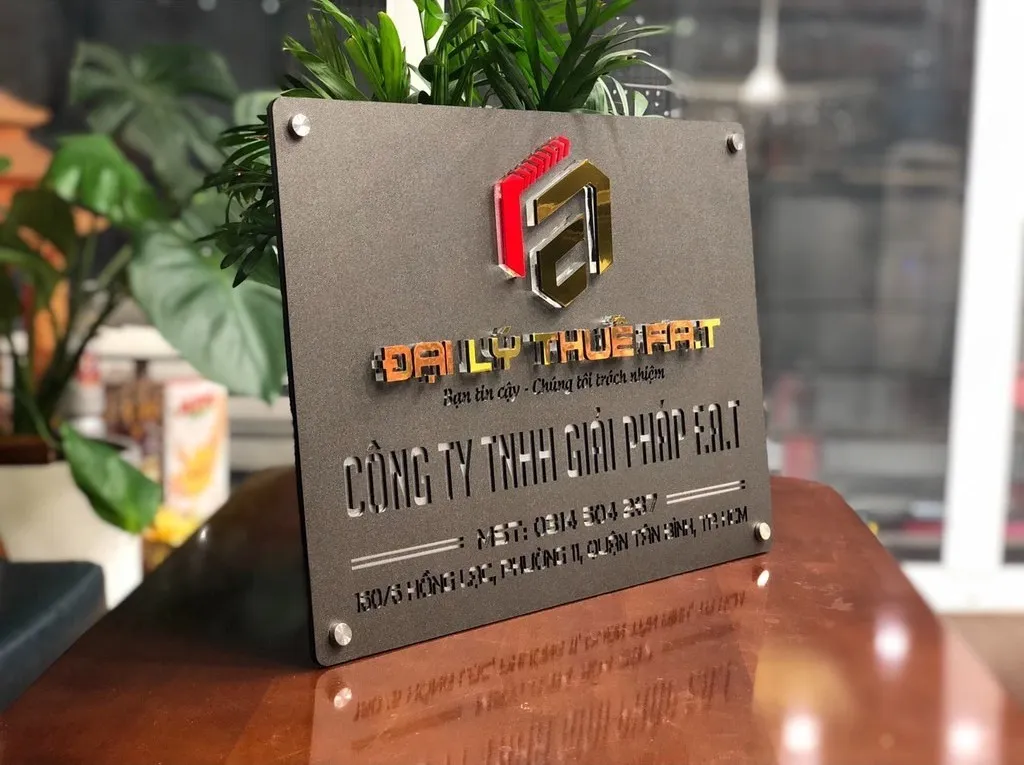 Bảng hiệu công ty inox xám khói logo 3D vàng đỏ bạc phối cảnh thực tế đặt trên bàn văn phòng cây xanh phía sau