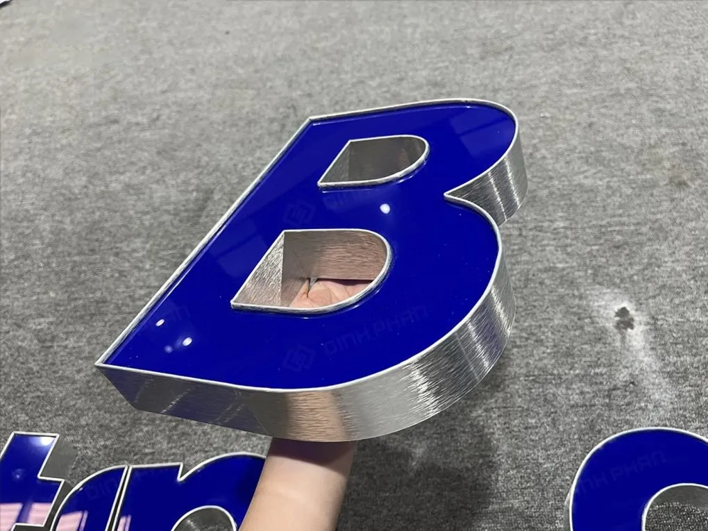 Cận cảnh chữ B thể hiện viền inox xước dày dặn và mặt mica xanh bóng