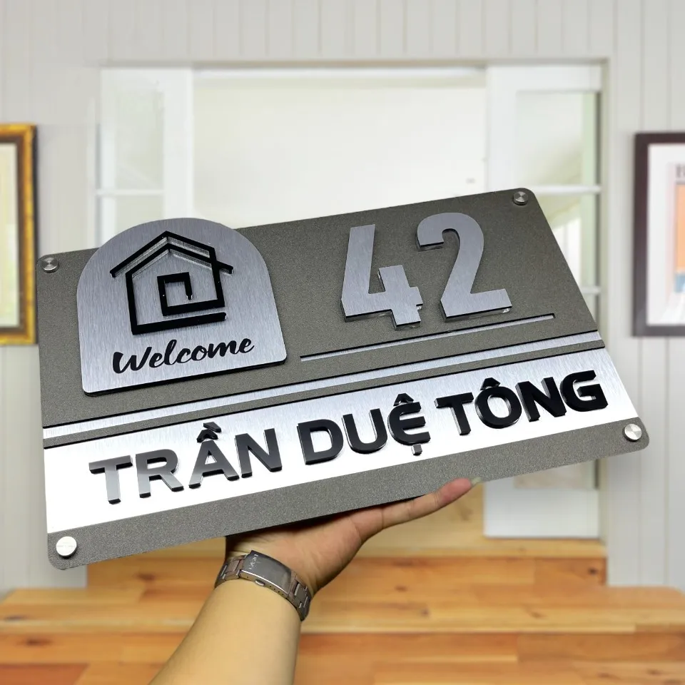 Close-up biểu tượng ngôi nhà inox đen cách điệu và chữ Welcome thủ bút trên bảng số nhà