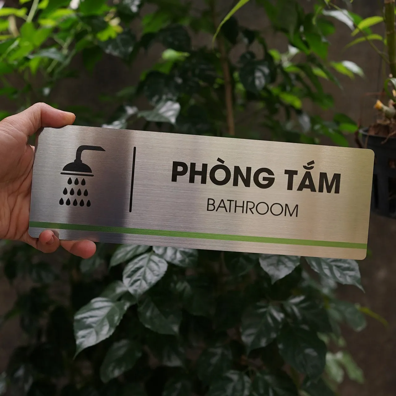 Bảng tên phòng tắm inox 304 phay xước cầm tay mặt trước nền cây xanh sáng