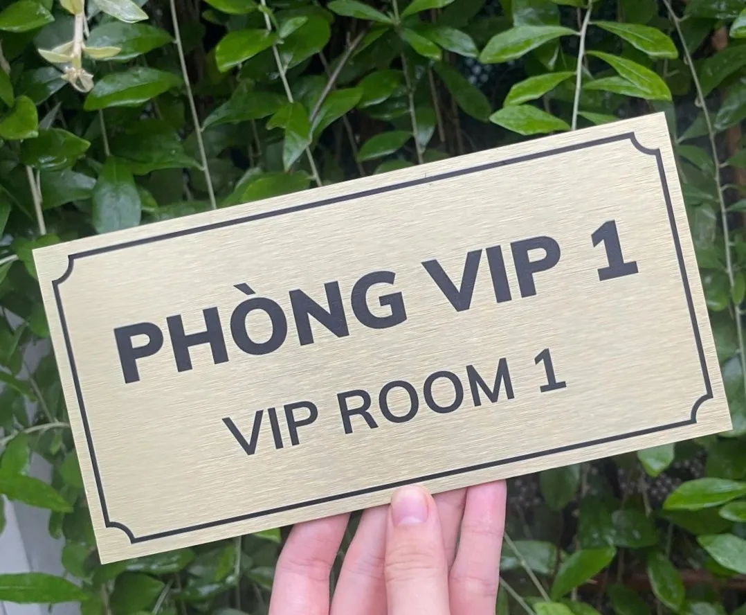 Bảng tên phòng VIP inox vàng xước nhìn từ góc nghiêng Bảng tên phòng VIP inox vàng xước cầm tay góc nghiêng nền lá xanh